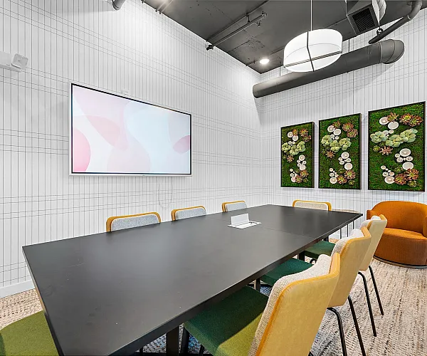 Contemporary meeting room featuring a sleek black conference table, vibrant chairs, wall-mounted screen, and artistic plant decor on textured walls.