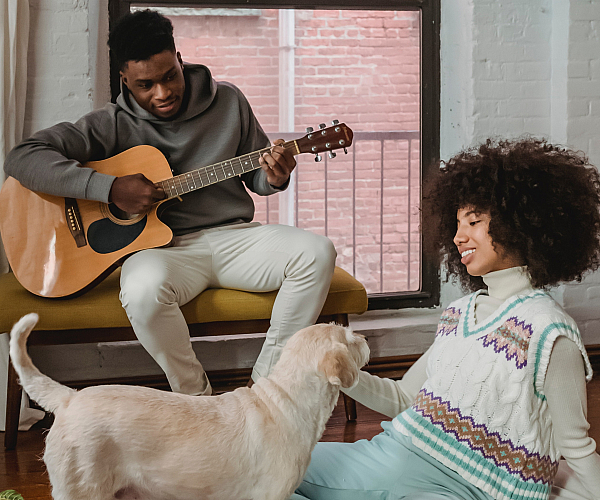 Cozy living space featuring a warm atmosphere with a person playing guitar, a relaxed individual seated on a patterned rug, and a friendly dog creating a welcoming vibe.