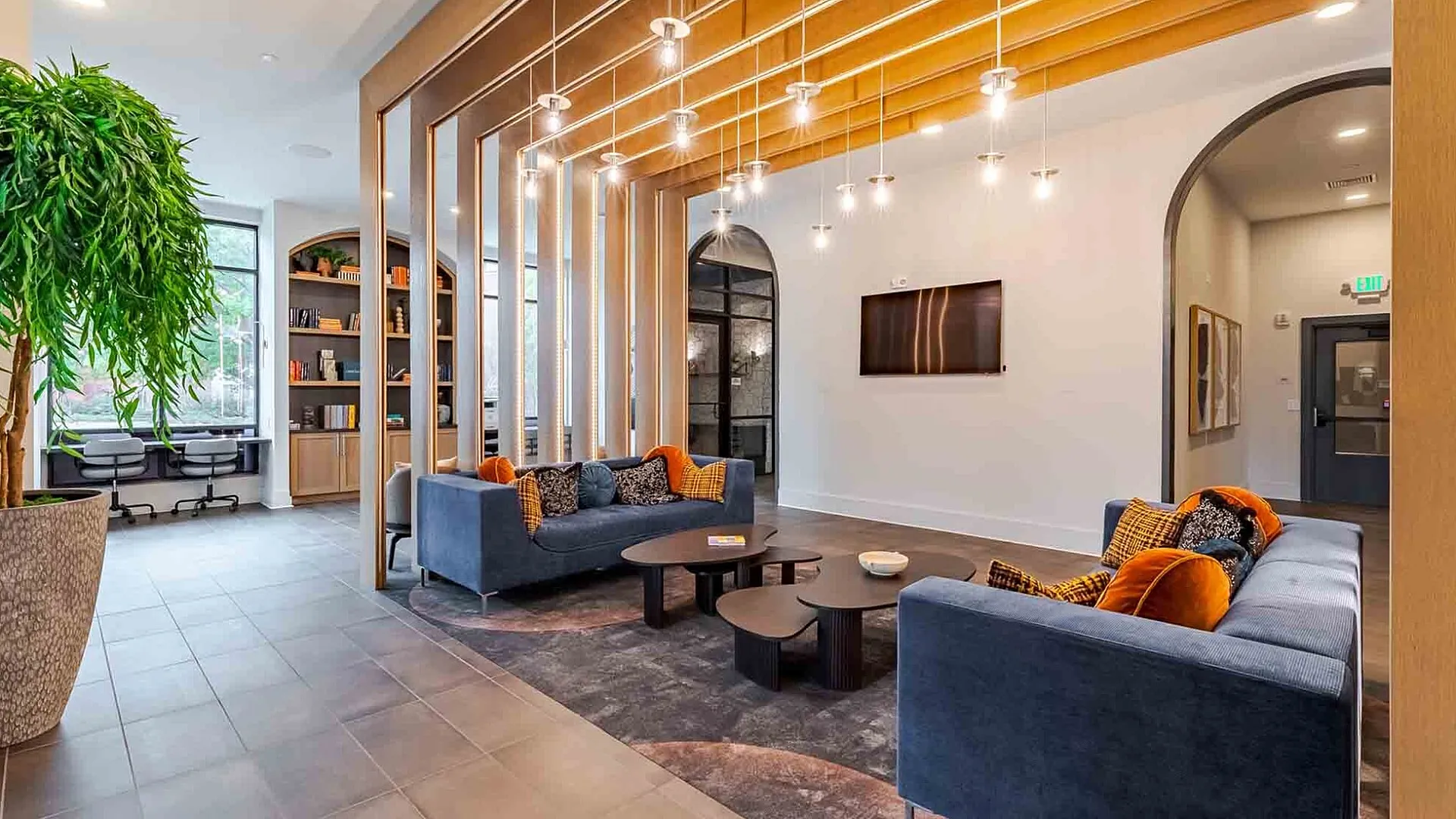Modern apartment lounge featuring stylish blue sofas, vibrant orange cushions, and sleek lighting. Includes a dining area, large plant, and decorative shelving with a cozy study nook.