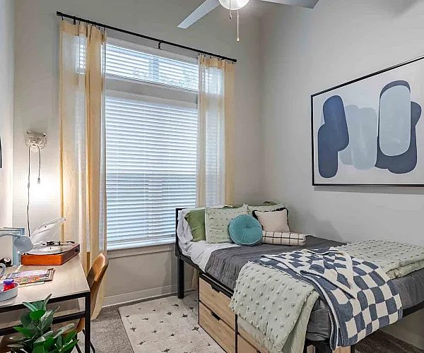 Cozy bedroom retreat with large window, stylish bedding, and a chic desk setup. Modern artwork and natural light create an inviting atmosphere.