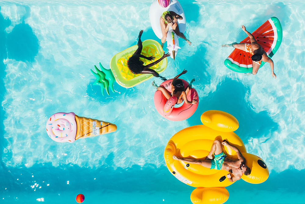 Vibrant aerial view of a sunny pool scene with colorful inflatable floats, including a unicorn, pineapple, watermelon, and ice cream cone, surrounded by people enjoying the water.