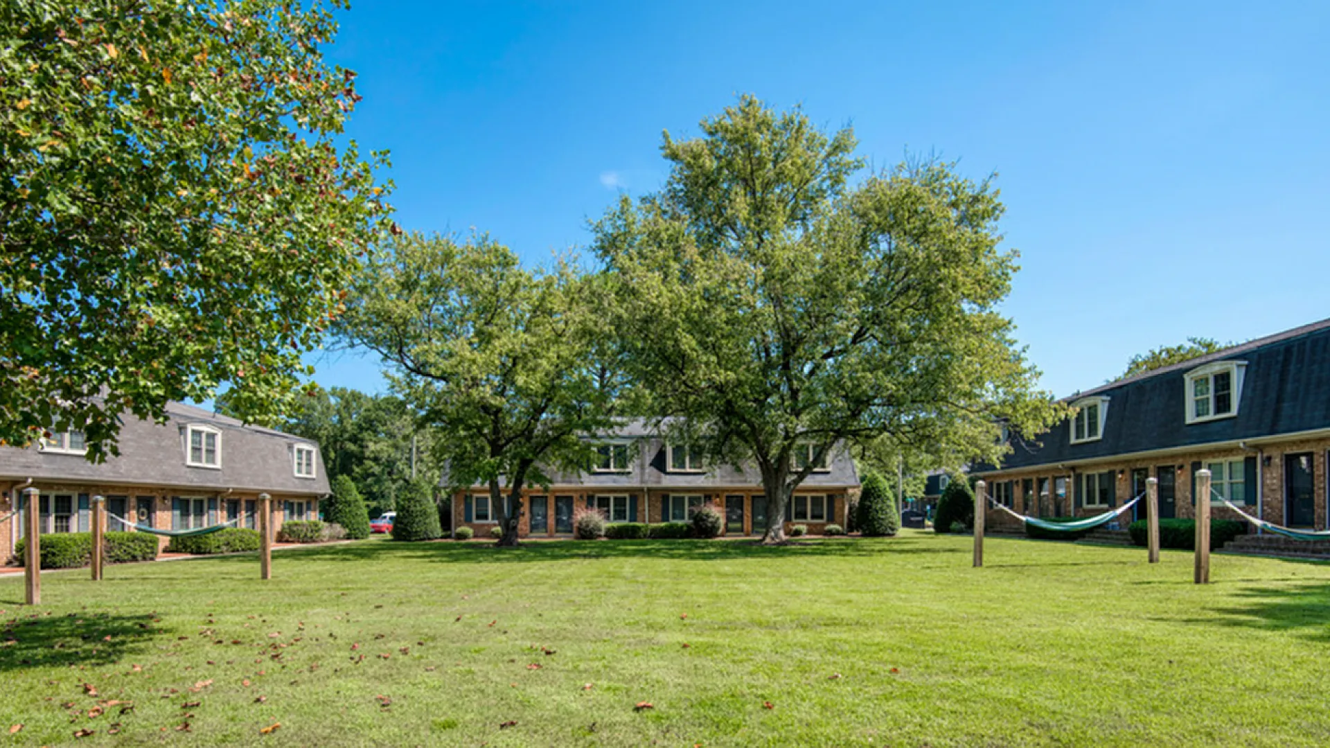 Charming apartment complex featuring brick facades, lush green lawns, and relaxing hammocks beneath mature trees on a sunny day.