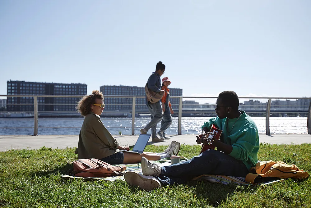 Spacious outdoor riverside area with people enjoying a sunny day; a person playing guitar and another using a laptop on the grass, with a stunning skyline and waterfront view.