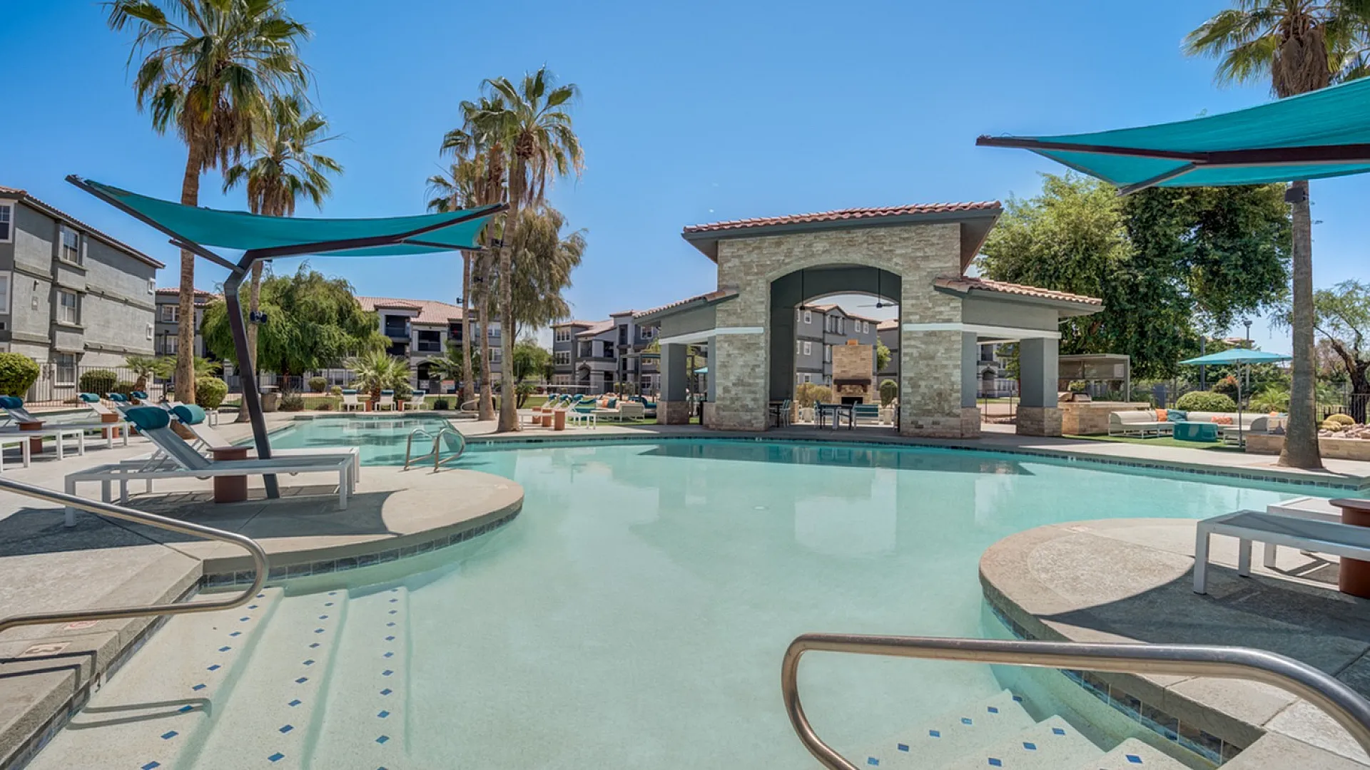 Luxurious apartment complex featuring a sparkling resort-style pool with stylish loungers and shaded canopies surrounded by lush palm trees.