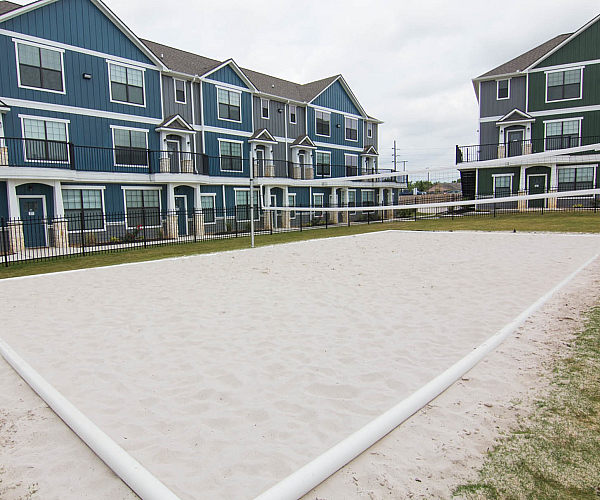Modern apartment buildings with elegant blue facades surround a pristine sand volleyball court, offering a vibrant community atmosphere.