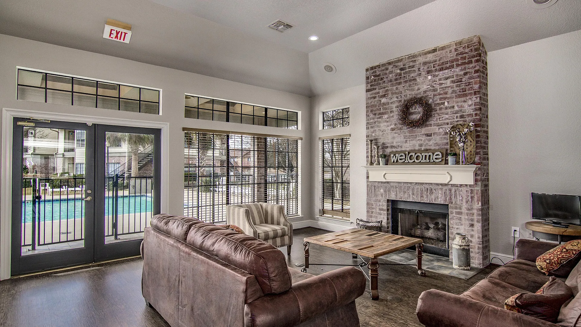 Inviting living room with plush brown sofas, a rustic brick fireplace, and large windows offering a view of the pool.