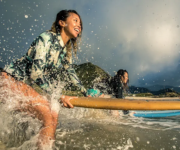 Exciting image of two women surfing, smiling as they ride the waves with a scenic mountain backdrop.
