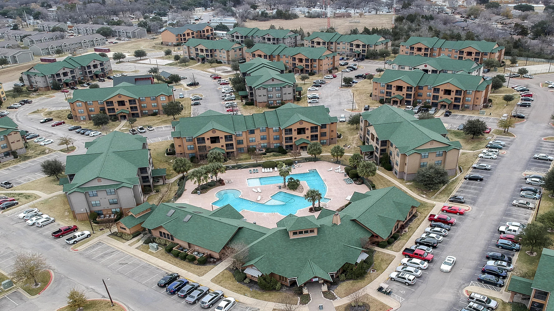 Aerial view of a charming apartment complex featuring multiple buildings with green roofs, ample parking, and a stylish central pool area.