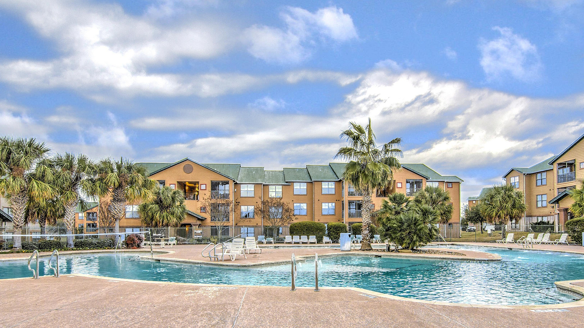 Inviting apartment complex featuring a sparkling pool surrounded by lush palm trees and cozy sun loungers, under a beautiful blue sky.