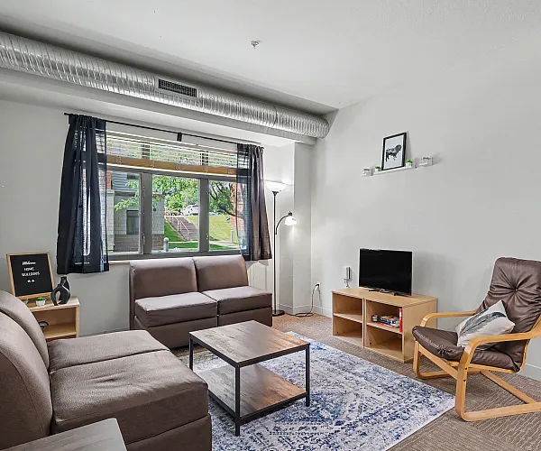 Inviting living room with modern decor, featuring cozy brown sofas, a sleek wooden coffee table, and a stylish armchair, enhanced by natural light from a large window with garden views.