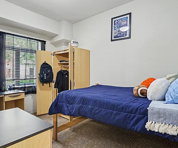 Cozy student bedroom with a neatly made bed, modern desk, and spacious wardrobe, bathed in natural light from a large window.