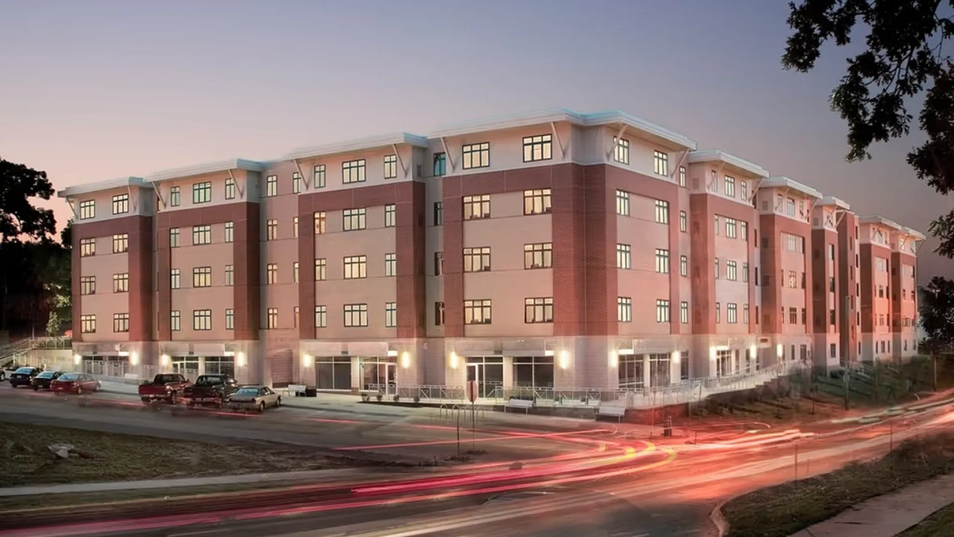 Charming five-story apartment building with modern architecture, illuminated by warm lights at twilight, featuring ample parking and a vibrant urban backdrop.