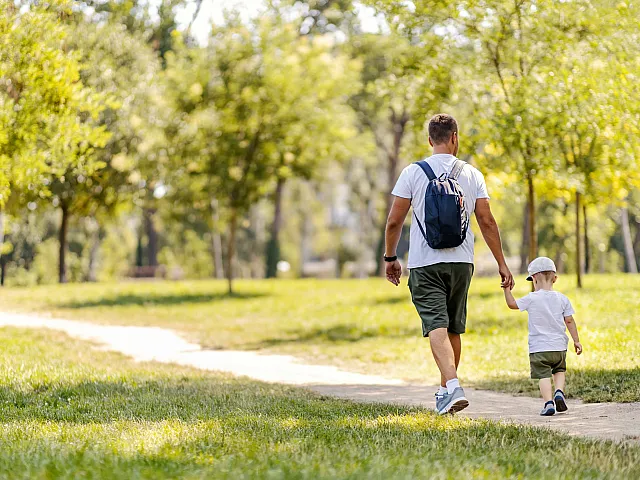 Charming park scene with a person and a child enjoying a serene walk on a sunny day, surrounded by lush greenery and trees.