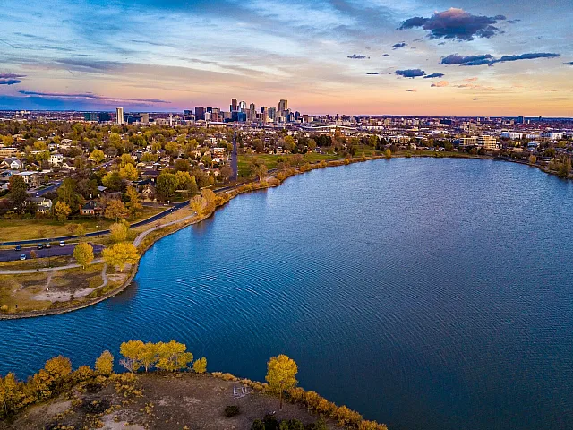 Breathtaking aerial view of a serene lake bordered by lush greenery and a vibrant city skyline at sunset.