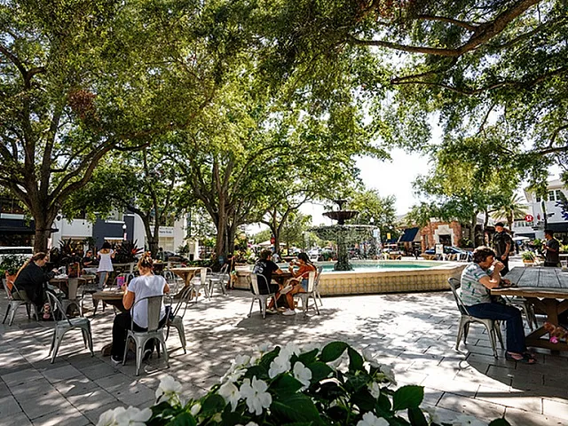 Charming outdoor plaza with people enjoying leisurely dining. Lush trees provide shade around a central fountain, offering a vibrant and inviting atmosphere.