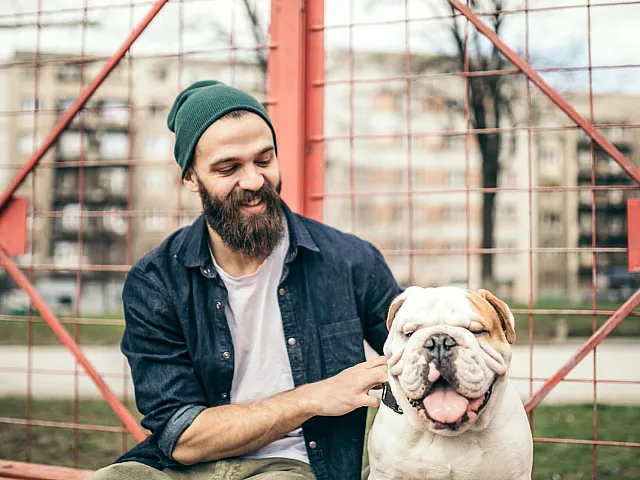 Cheerful man in a beanie joyfully petting a happy bulldog in a vibrant urban outdoor setting.