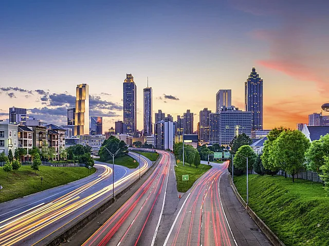 Breathtaking city skyline view with vibrant sunset over modern high-rises, surrounded by lush greenery and well-lit highways.