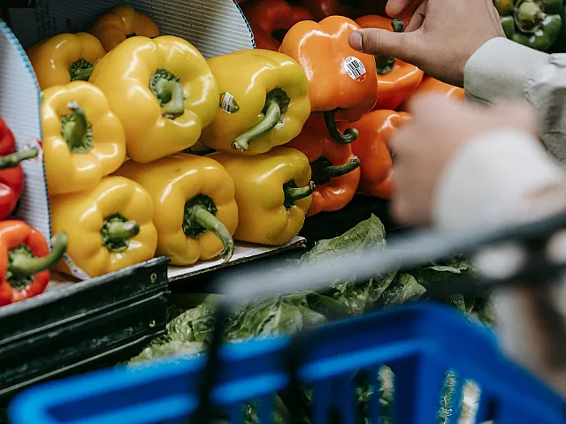 Vibrant grocery display featuring fresh yellow and orange peppers being selected by a shopper with a blue basket.