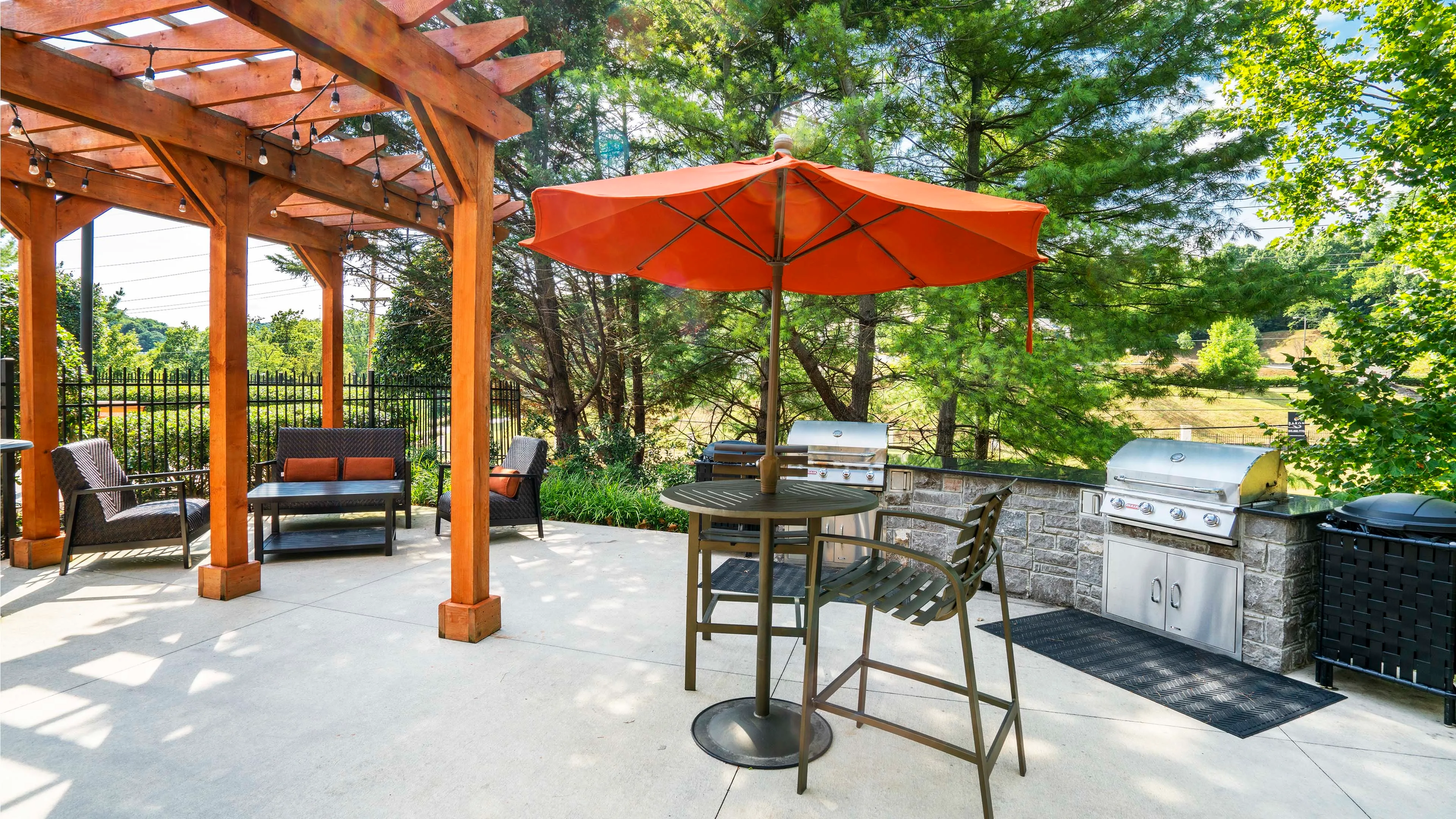 Inviting outdoor patio with a pergola, stylish seating, and a vibrant orange umbrella, perfect for entertaining by the grill amidst lush greenery.
