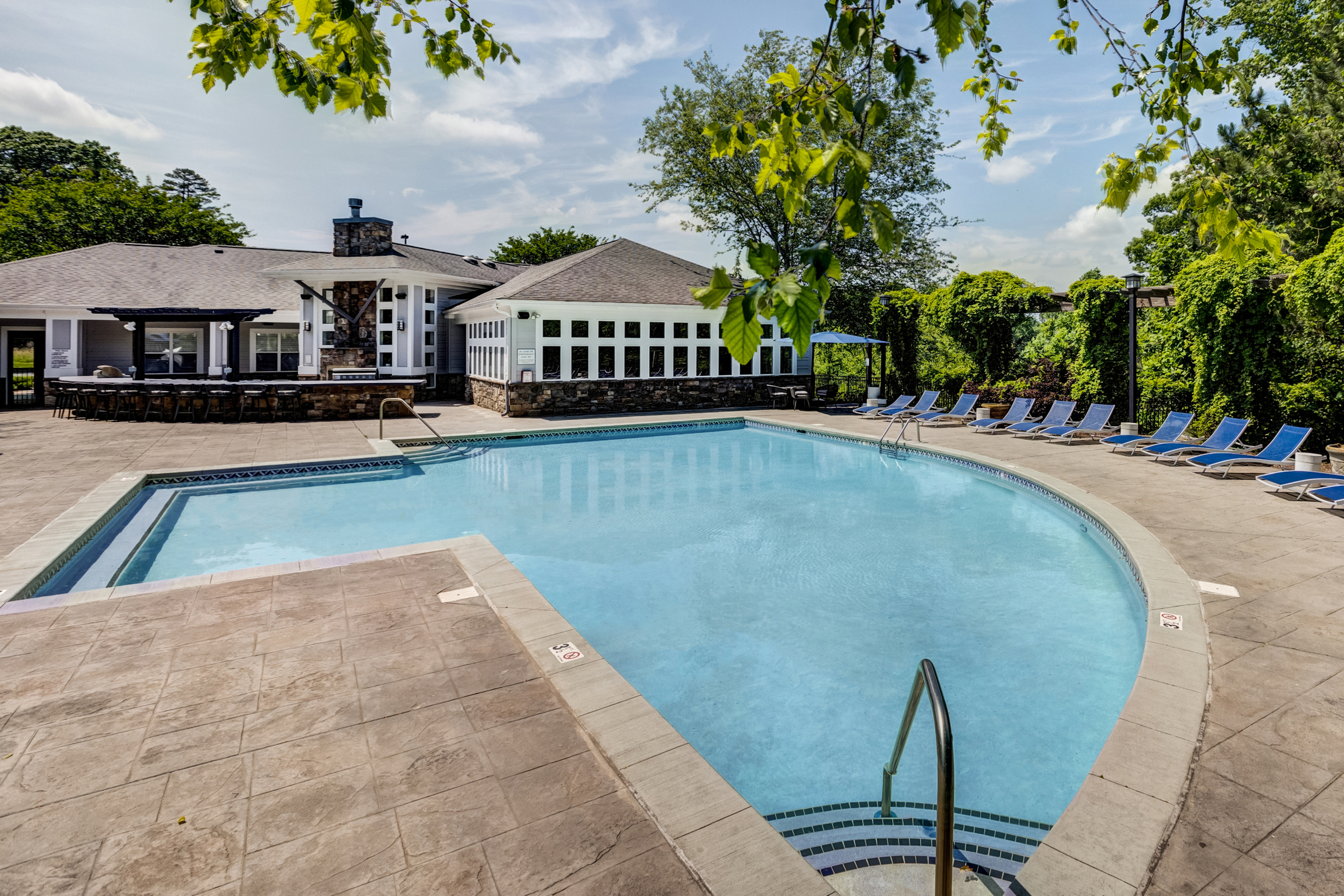 Inviting apartment complex pool area with sun loungers, surrounded by lush greenery and a charming clubhouse.