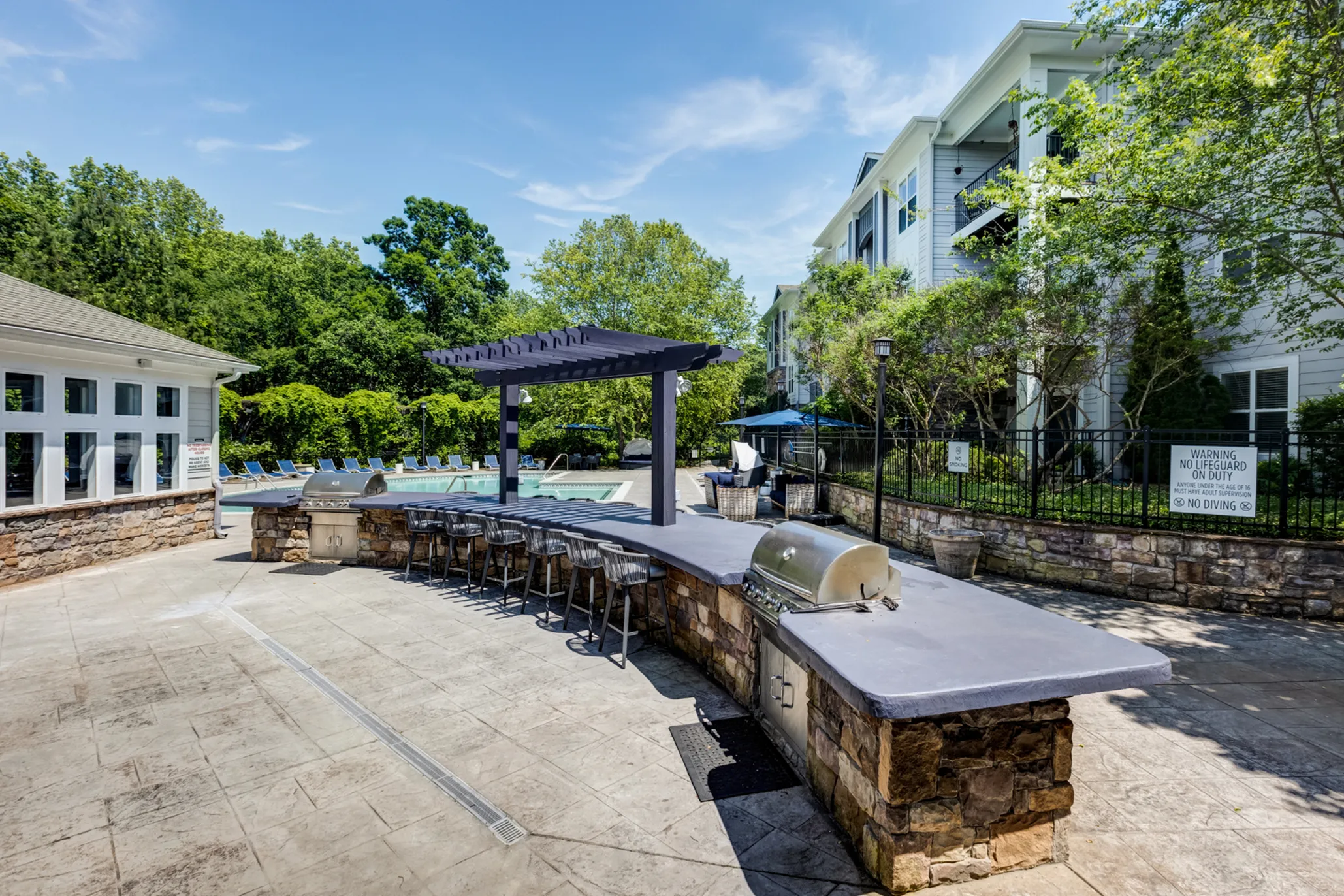 Inviting outdoor area with a modern grill station, bar seating under a stylish pergola, and a serene swimming pool surrounded by lush greenery.
