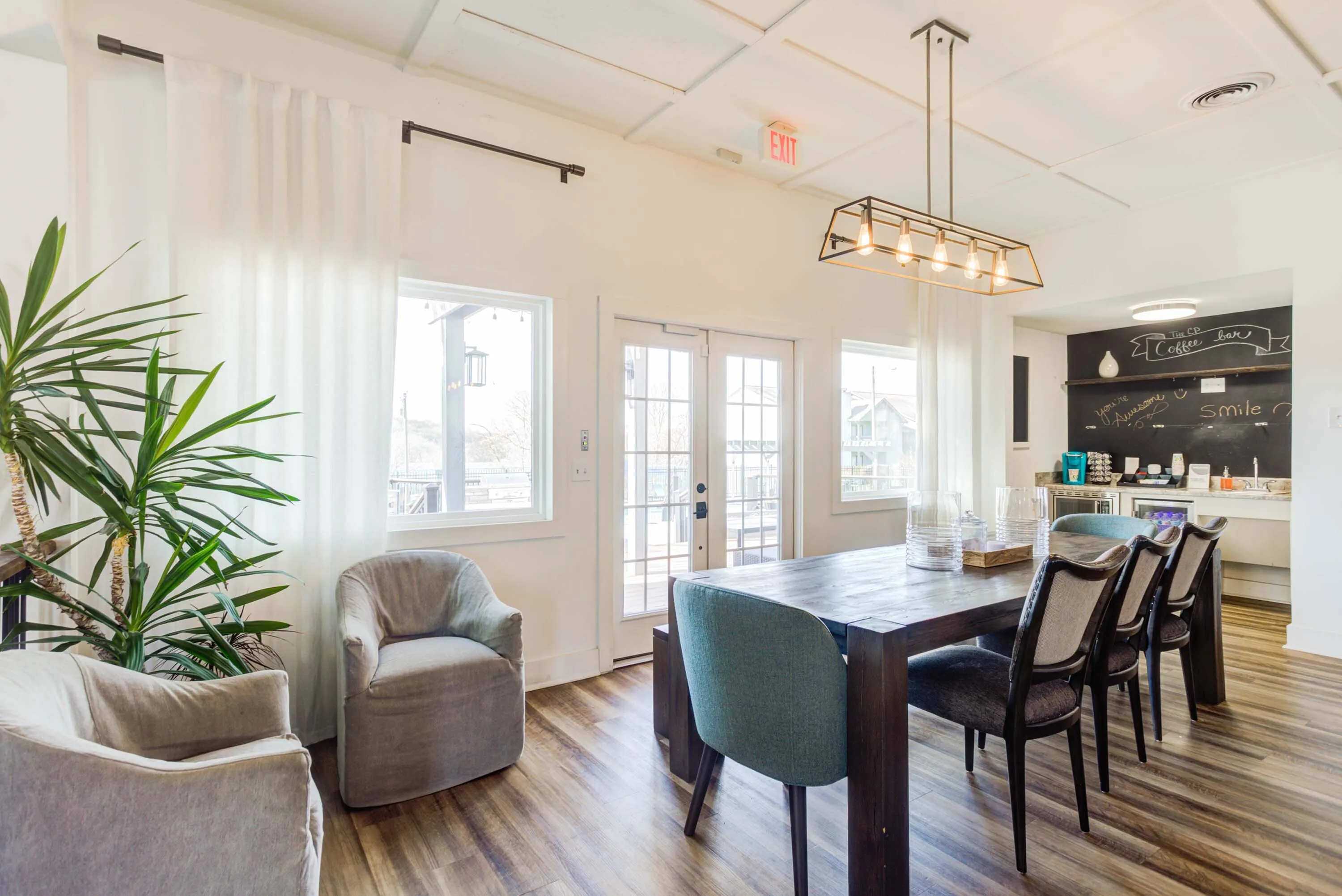 Bright, modern dining room with elegant lighting, large wooden table, and cozy seating. Natural light streams through windows, highlighting the sleek coffee bar in the background.