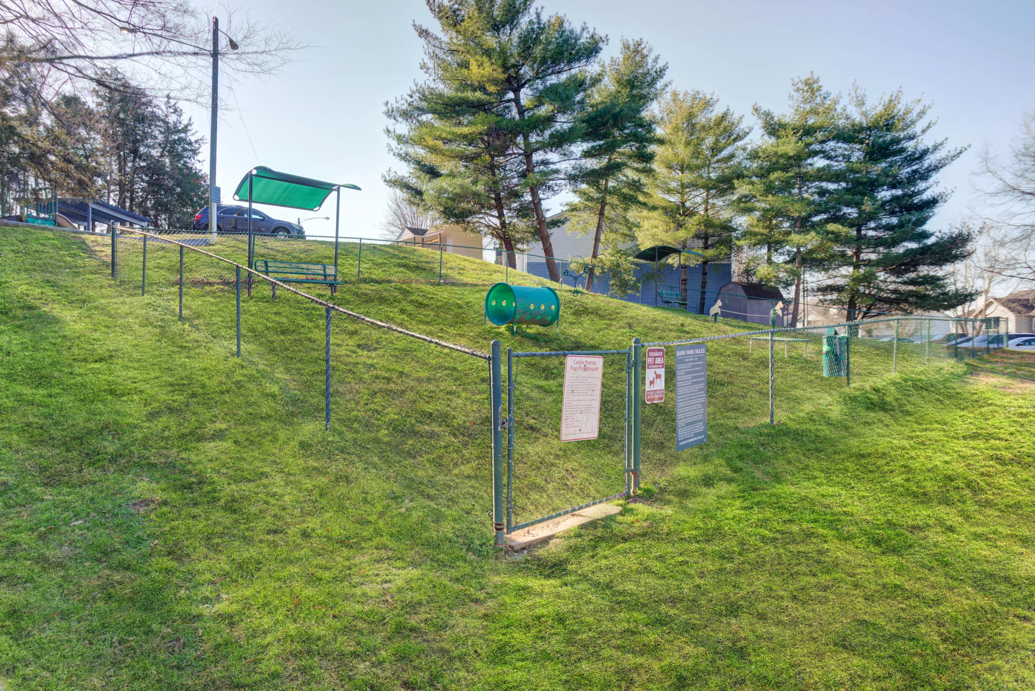 Charming dog park with lush green grass, shaded seating areas, and a playful tunnel under sunny skies.