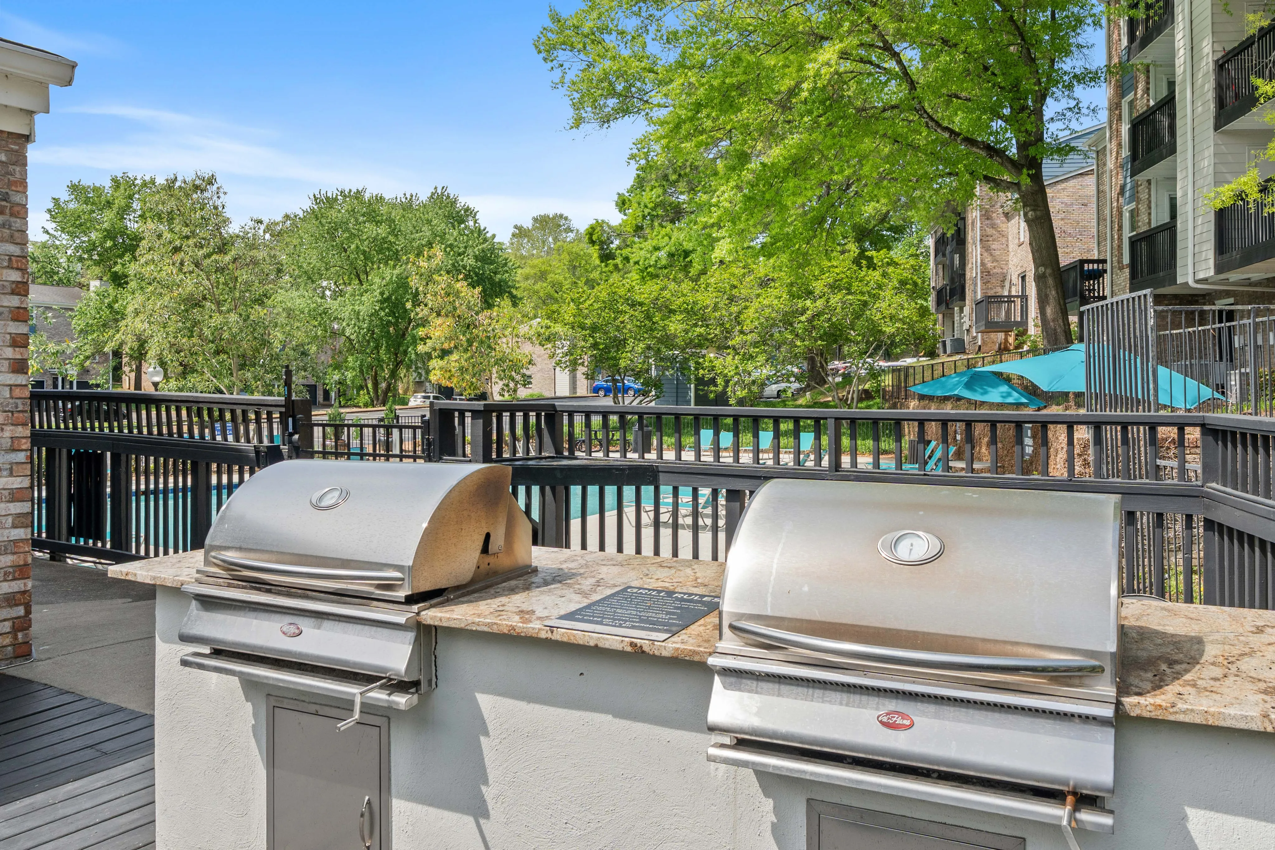 Inviting outdoor space featuring stainless steel grills and a view of a sparkling pool surrounded by lush greenery and modern apartments.