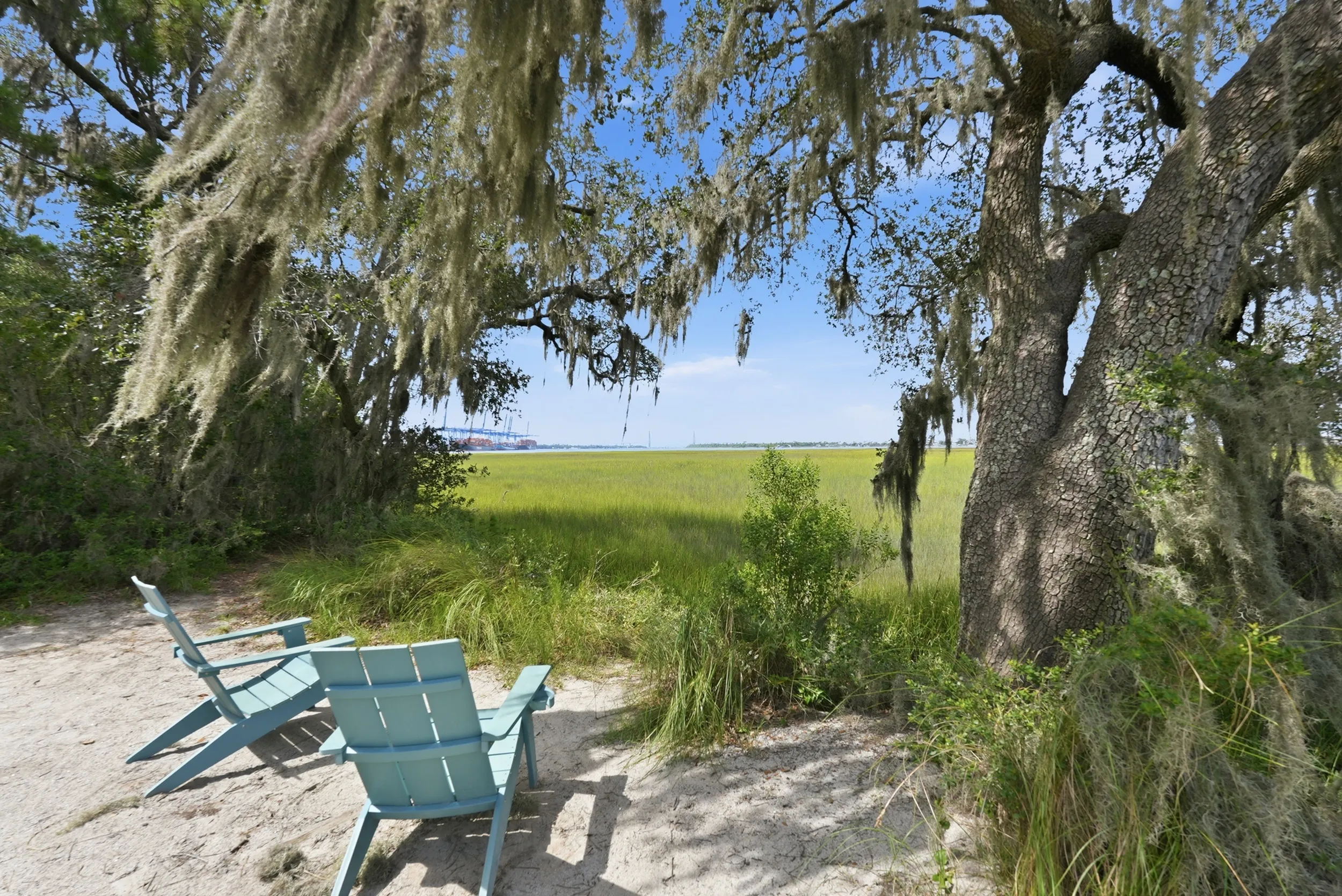 Scenic outdoor view featuring tranquil marshlands, a shady tree covered with Spanish moss, and two inviting Adirondack chairs.