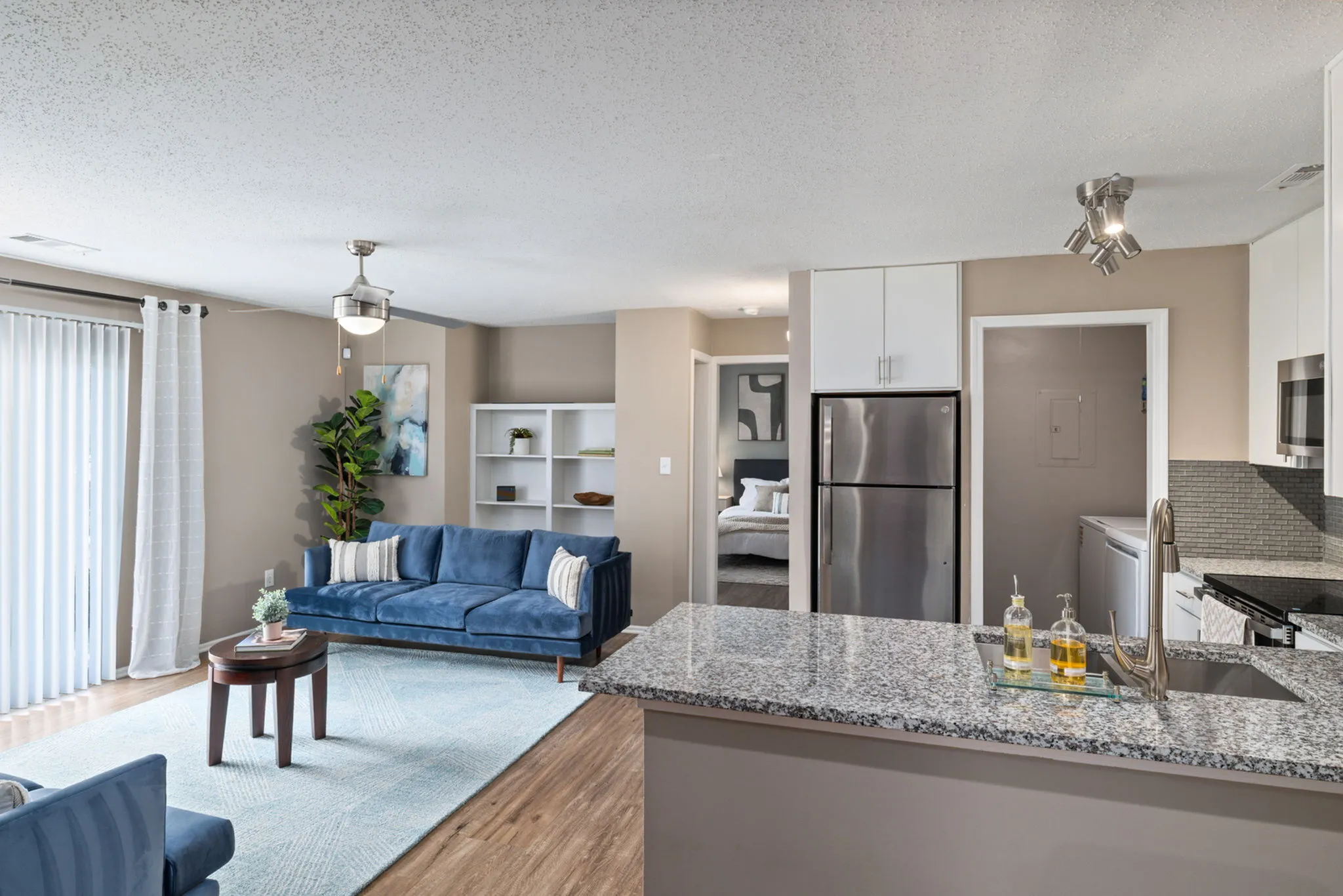 Contemporary open-plan living room with elegant blue sofas, modern kitchen, and sleek design, leading to a cozy bedroom.