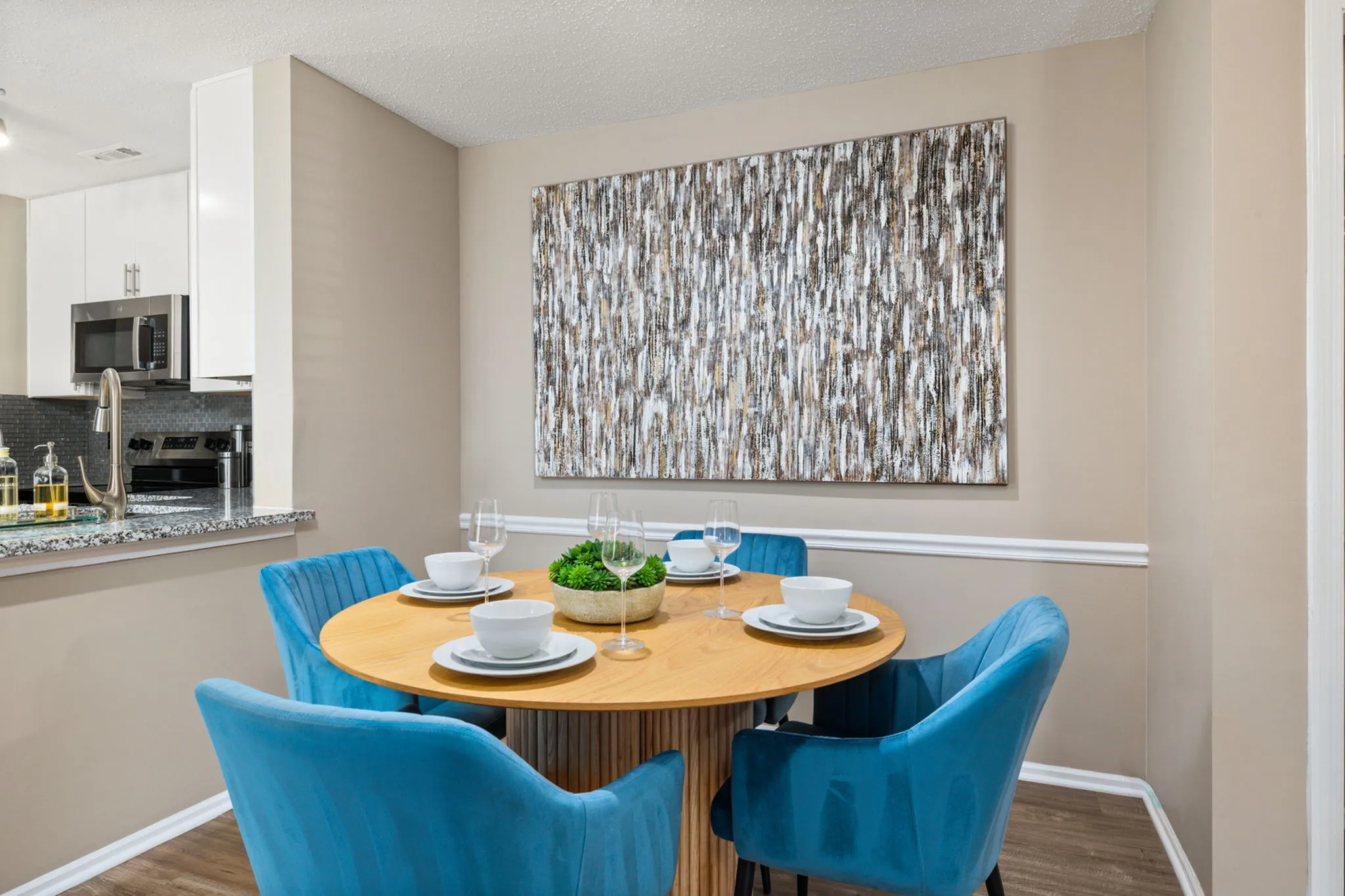 Contemporary dining area with a stylish round table set for four, featuring vibrant blue chairs and modern wall art, adjacent to a sleek kitchen with stainless steel appliances.