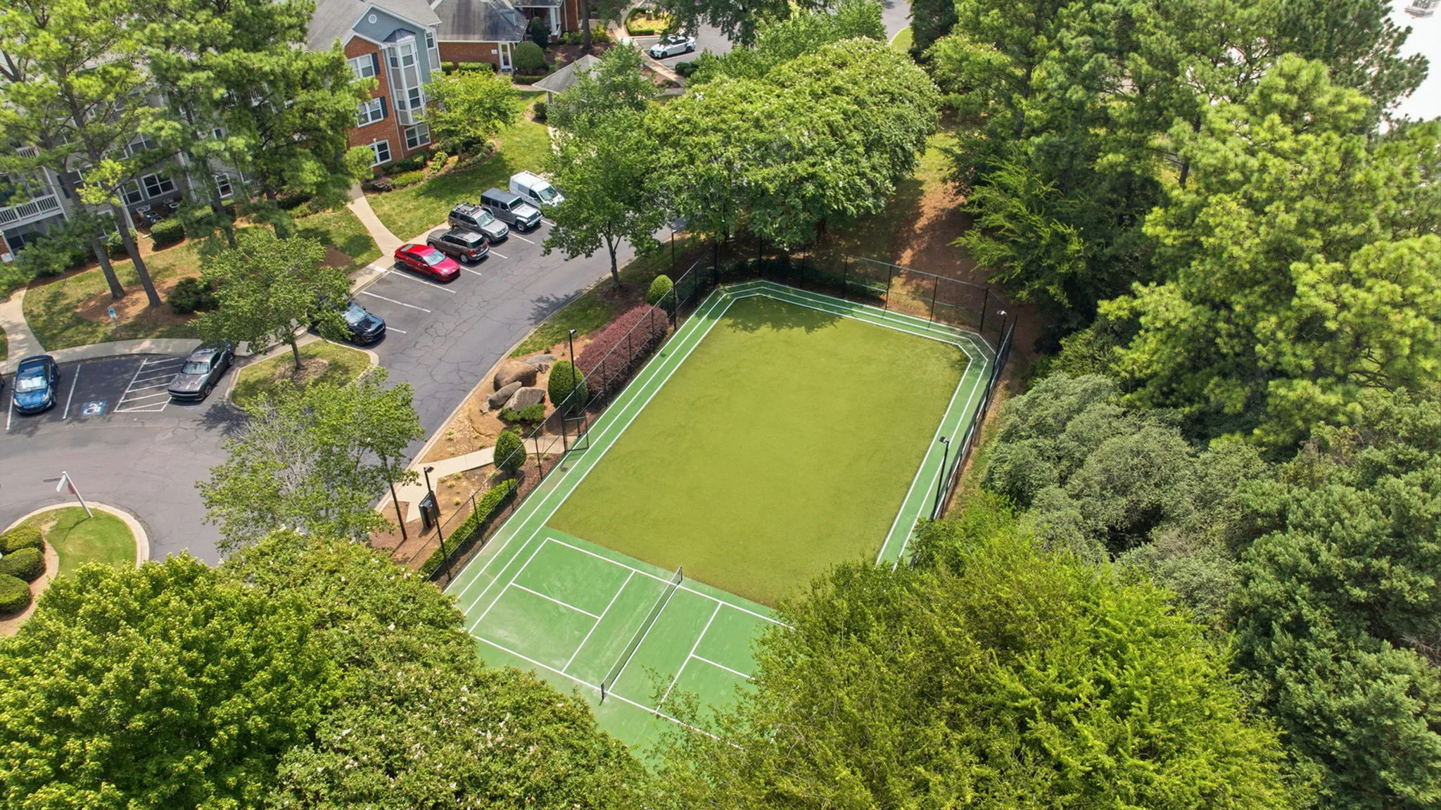 Aerial view of a delightful tennis court nestled amidst lush greenery, with apartments and parking area in the background, showcasing a serene residential community setting.