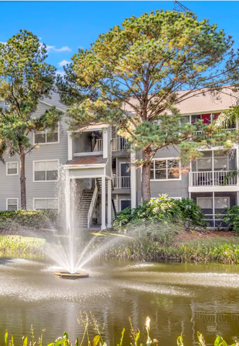 Exterior view of Avana City Park apartments in Clearwater, FL, featuring trees and a pond with a fountain.