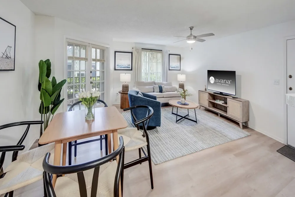 Model living room and dining space at Avana Coral Springs apartments, featuring wood grain flooring and large glass doors.
