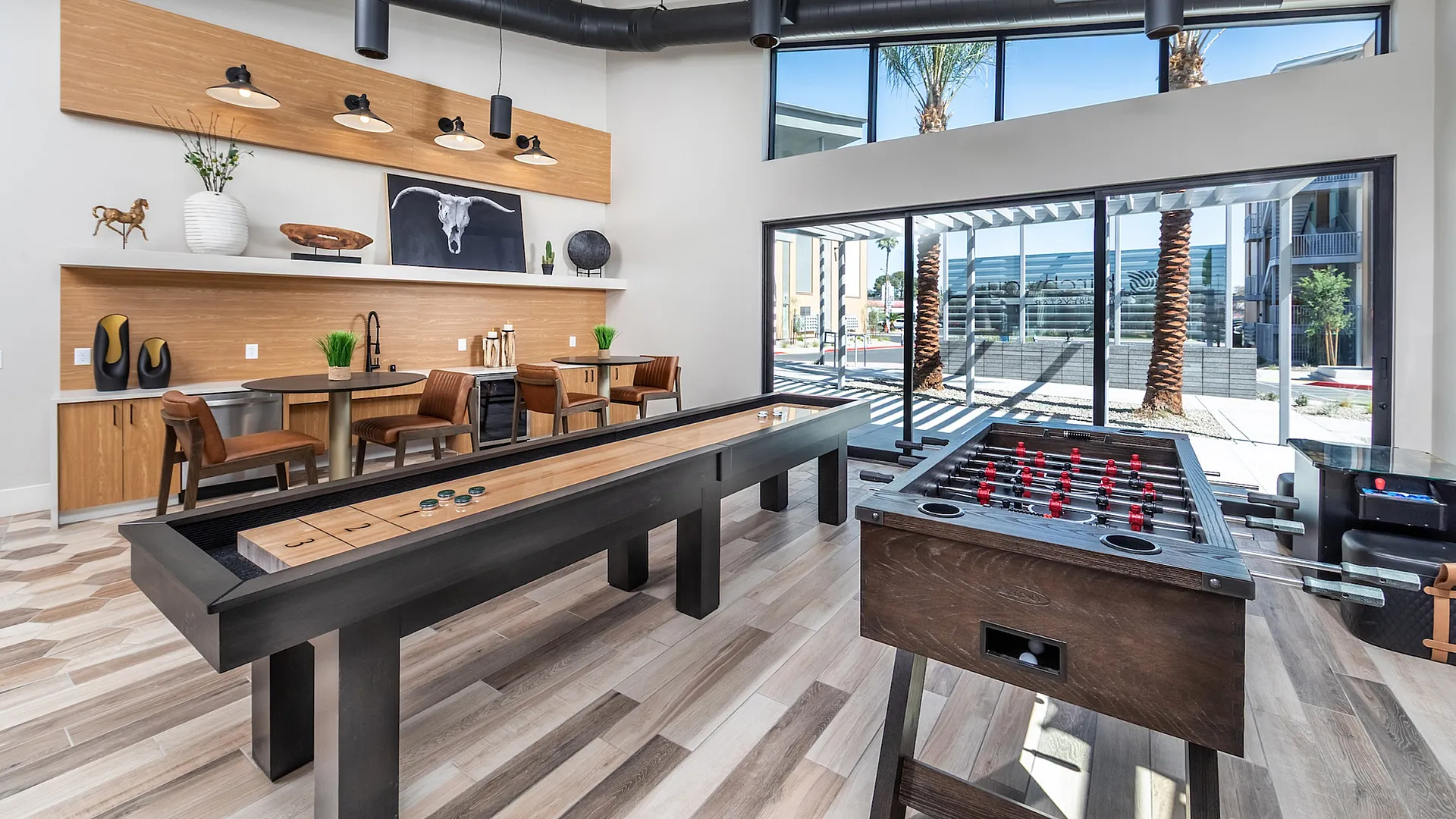Modern recreational room with sleek shuffleboard and foosball tables, ample seating, and stylish decor, accented by large windows offering scenic outdoor views.