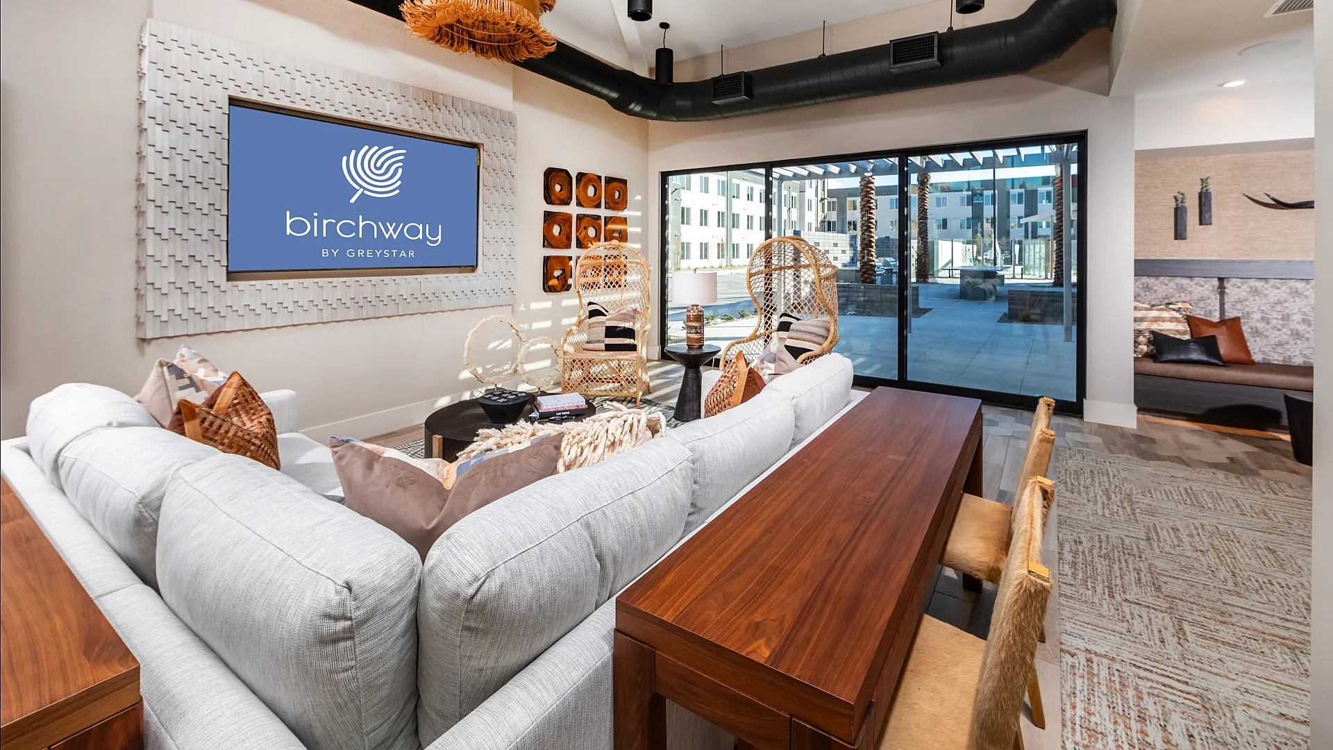 Modern apartment lounge with elegant decor, featuring plush sofas, wicker chairs, and a large window offering a vibrant urban view.