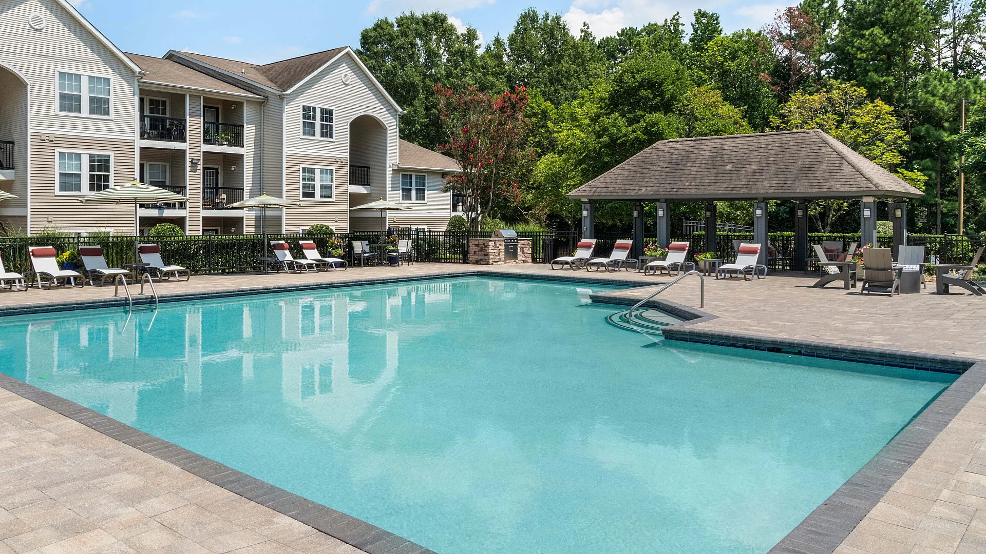 Inviting apartment complex with a crystal-clear swimming pool, surrounded by elegant lounge chairs and lush greenery, offering a serene escape.