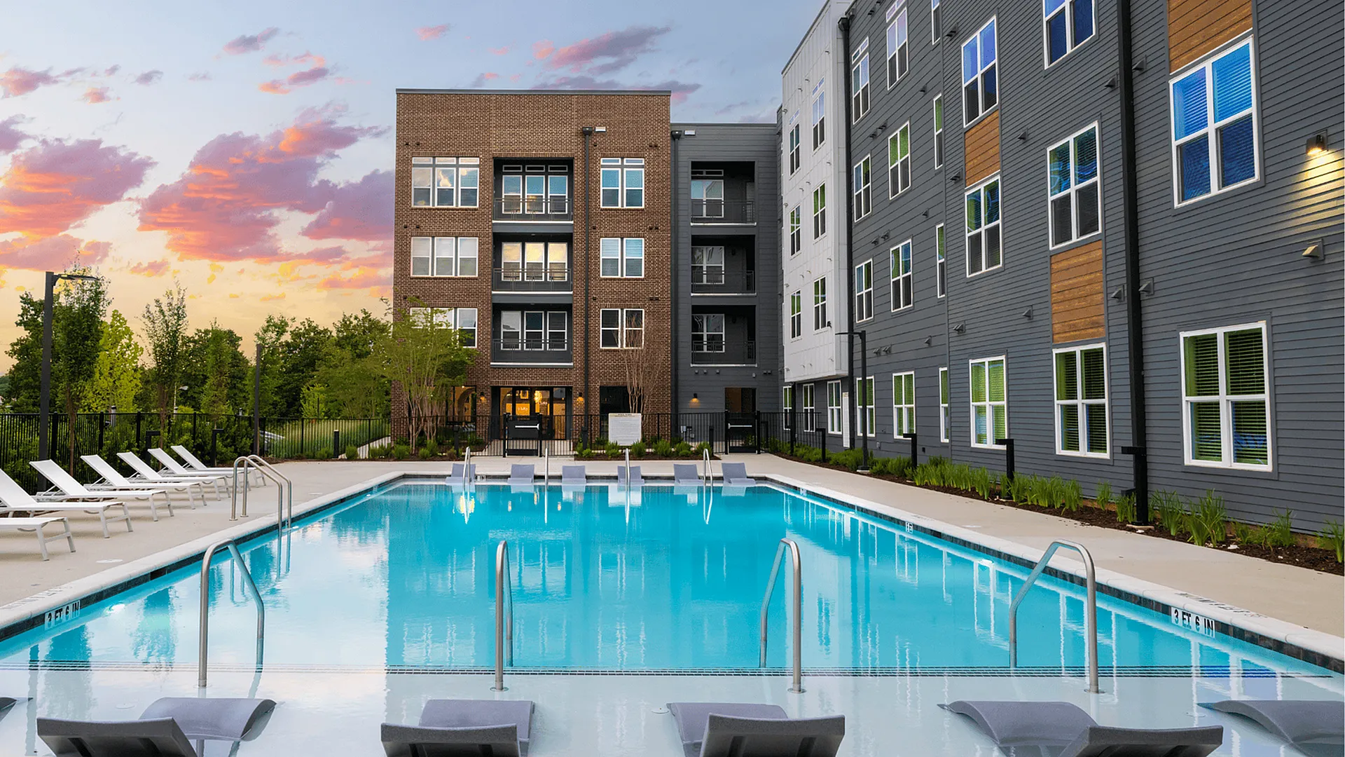 Modern apartment complex featuring a sparkling outdoor pool with lounge chairs, surrounded by lush greenery and stunning architecture against a vibrant sunset sky.