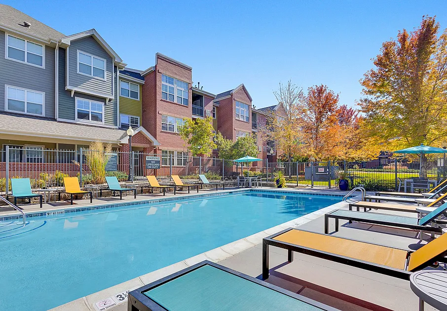 Inviting apartment complex with a sparkling outdoor pool, colorful loungers, and vibrant autumn trees, offering a perfect blend of relaxation and modern living.