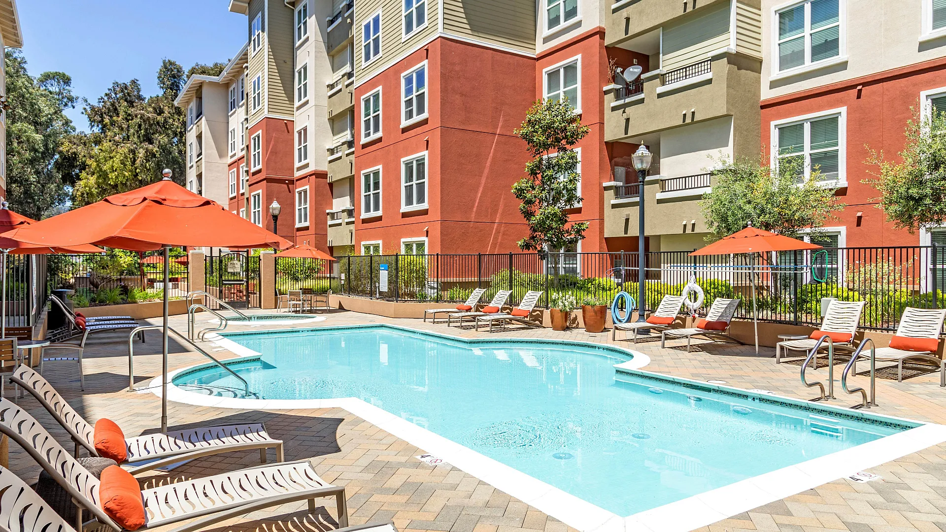 Inviting apartment complex with a sunlit pool area, vibrant red and beige architecture, and comfortable poolside loungers with orange umbrellas.