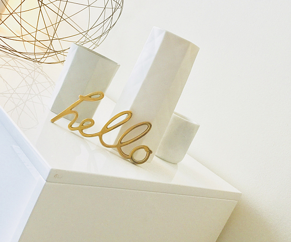 Contemporary interior decor featuring a sleek white surface, elegant geometric wire sculpture, and stylish 'hello' lettering in gold.