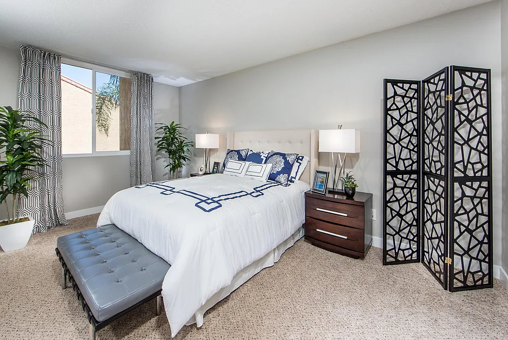 Elegant bedroom with cozy beige carpet, stylish patterned curtains, plush bed with crisp white linens, chic wooden nightstands, and lush green plants enhancing the serene ambiance.