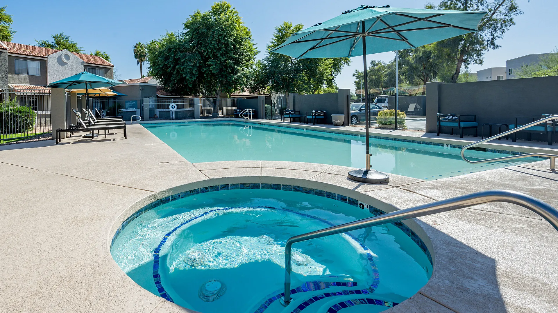 Inviting swimming pool area with comfortable lounge chairs and sun umbrellas, surrounded by lush greenery and modern apartment buildings.