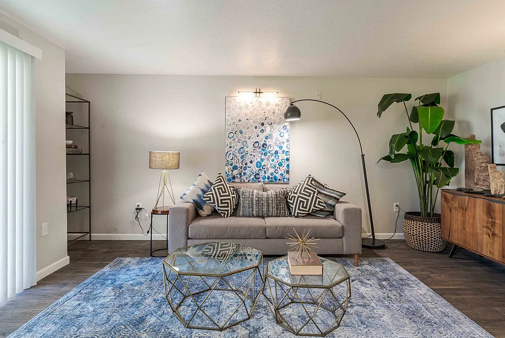 Chic and inviting living room featuring a contemporary gray sofa adorned with geometric cushions, stylish glass coffee tables, and a plush blue rug, enhanced by elegant lighting and lush greenery.