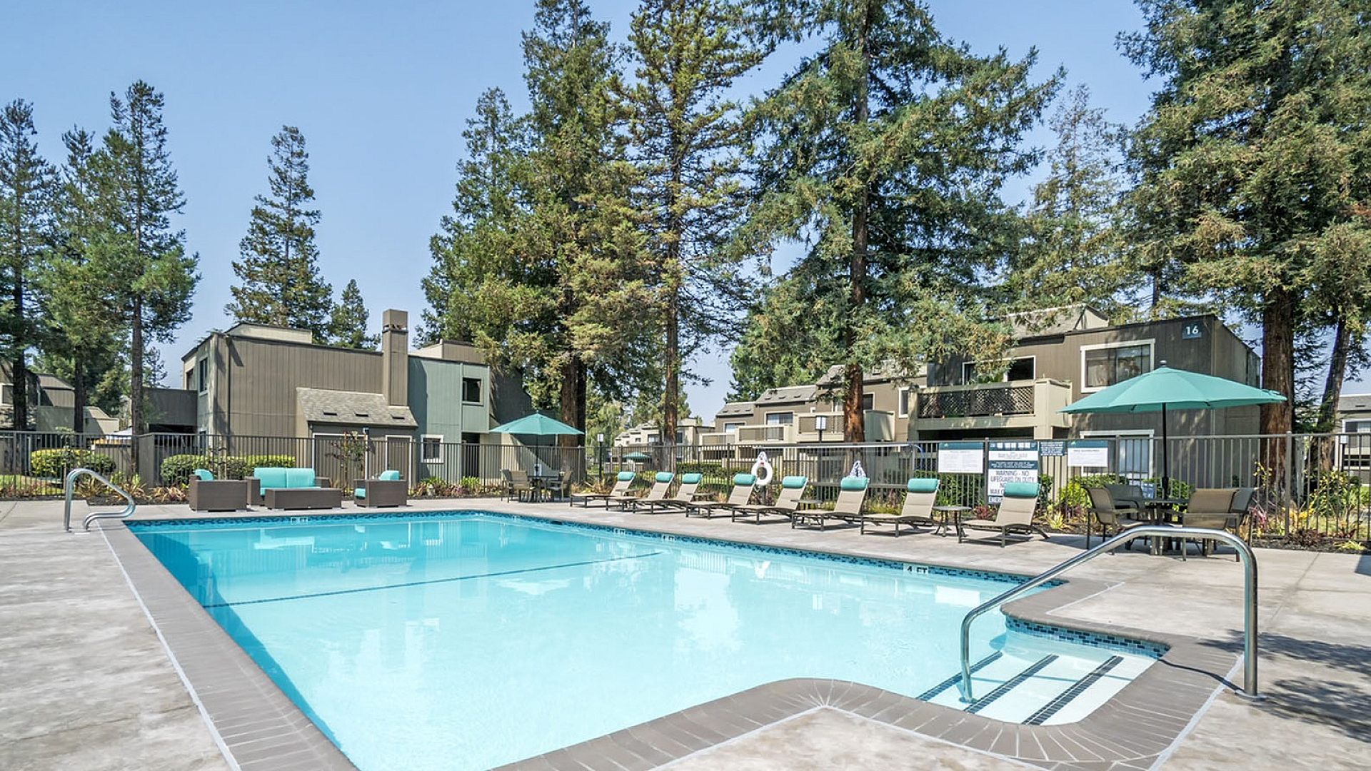 Inviting outdoor pool area with sleek lounge chairs and umbrellas, surrounded by lush pine trees in a serene residential setting.