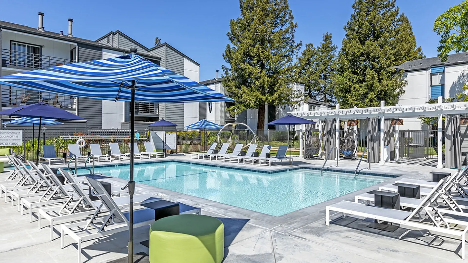 Inviting apartment complex pool with stylish lounge chairs, vibrant umbrellas, and lush greenery, ideal for relaxing in sunny weather.
