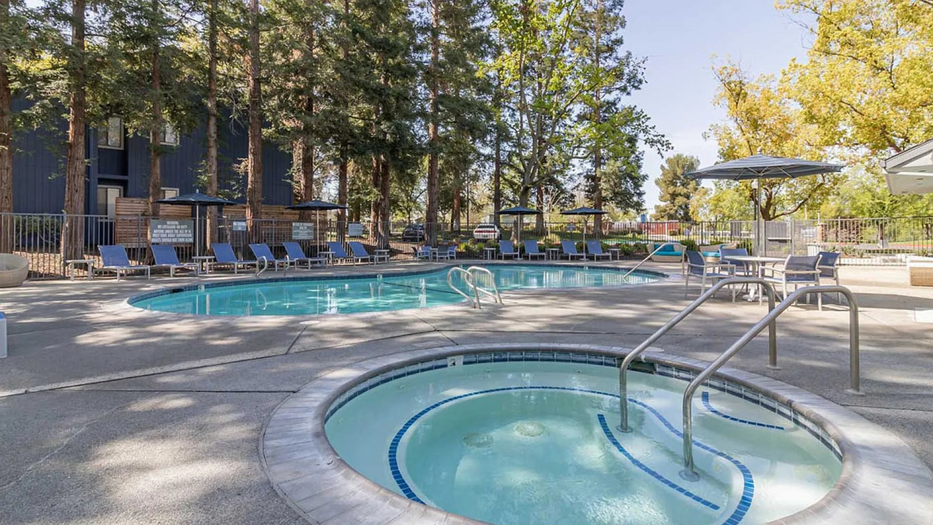 Inviting outdoor pool area with a sunlit hot tub, surrounded by lush trees and comfortable lounge chairs, perfect for relaxation.