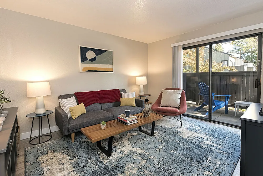 Inviting living room with modern decor, featuring a cozy gray sofa, stylish coffee table, and bright lighting. Glass sliding doors lead to a charming patio with outdoor seating, perfect for relaxation.
