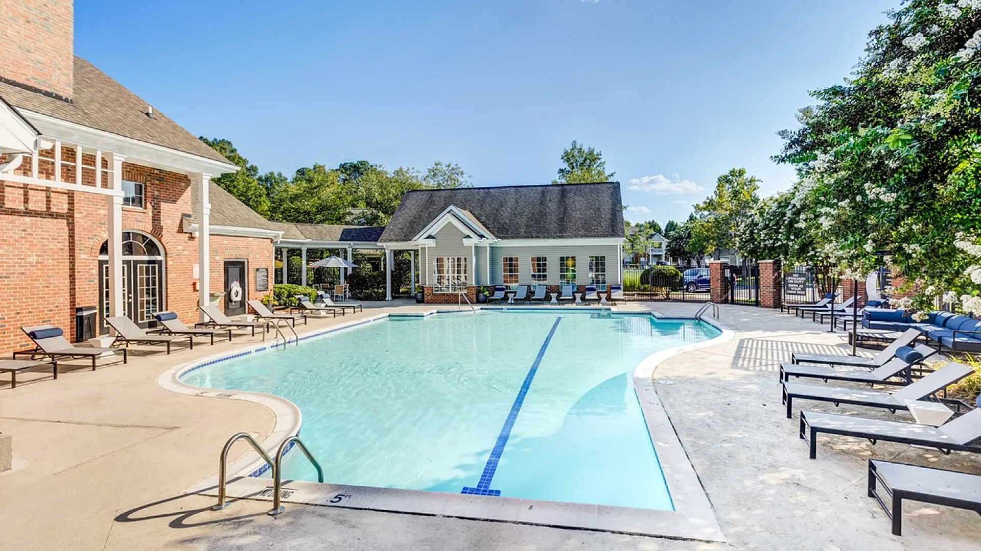 Luxurious outdoor pool area with elegant lounge chairs and a charming clubhouse, surrounded by lush greenery and a clear blue sky.