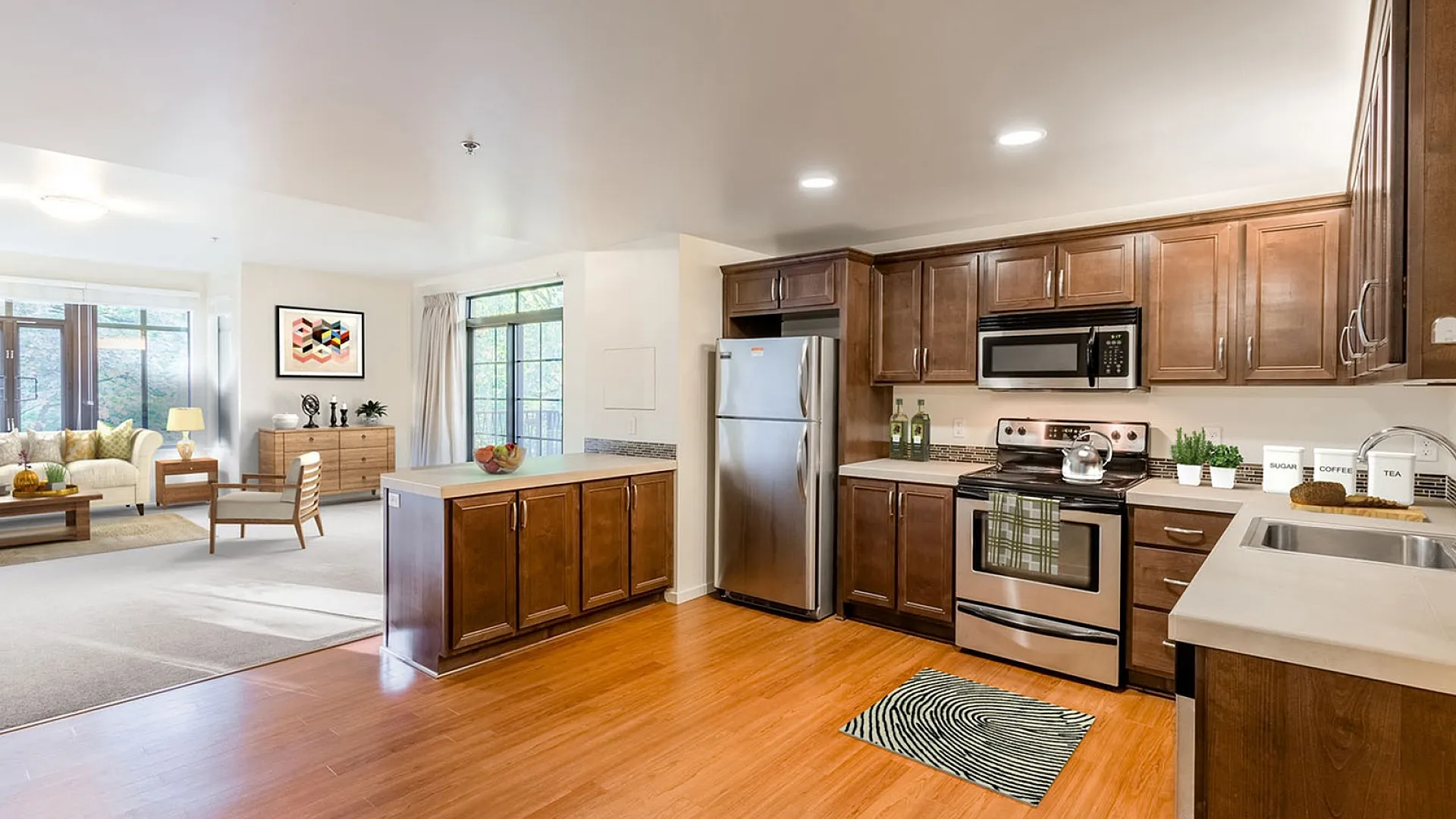 Modern open-concept living and kitchen area with sleek appliances, warm wooden cabinets, and abundant natural light from large windows.